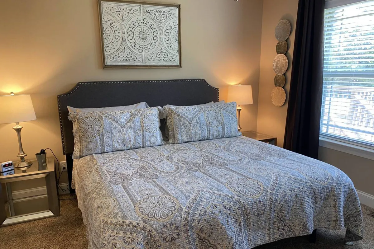 A bright, inviting bedroom with a plush bed, matching bedside lamps, attractive wall art, and heavy curtains. Sunlight gently filters through the window, illuminating the soft linens and creating a cozy, restful atmosphere.