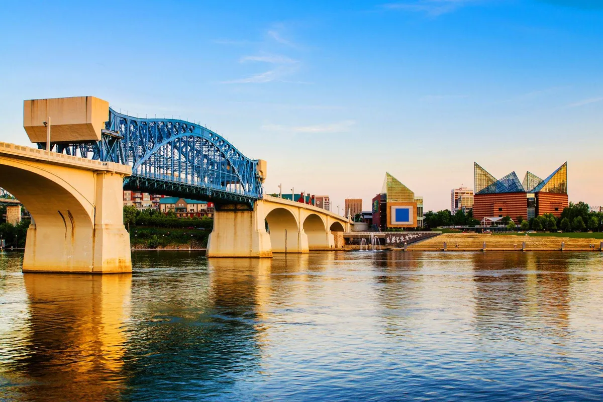 A scenic view of a large bridge spanning water, with buildings in the background and an expansive sky overhead. The image hints at the Tennessee Riverwalk and the blend of urban and natural appeal found in Chattanooga.