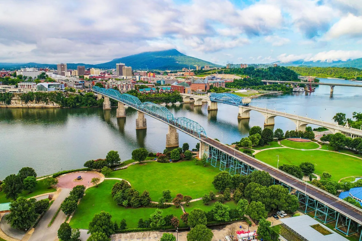 A sweeping view of the river bordered by city bridges and the Chattanooga skyline with mountains in the distance. The waterfront is lively, suggesting opportunities for walking, cycling, and outdoor exploration.