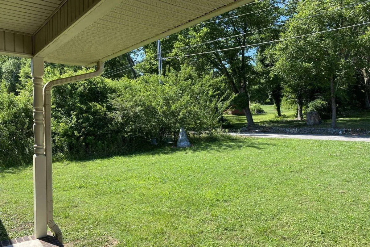 A scenic exterior of the property with lush green grass, mature trees, and a spacious porch area. The image conveys a calm and serene mood, perfect for hosting small gatherings, relaxing outdoors, or reflecting after a big event.
