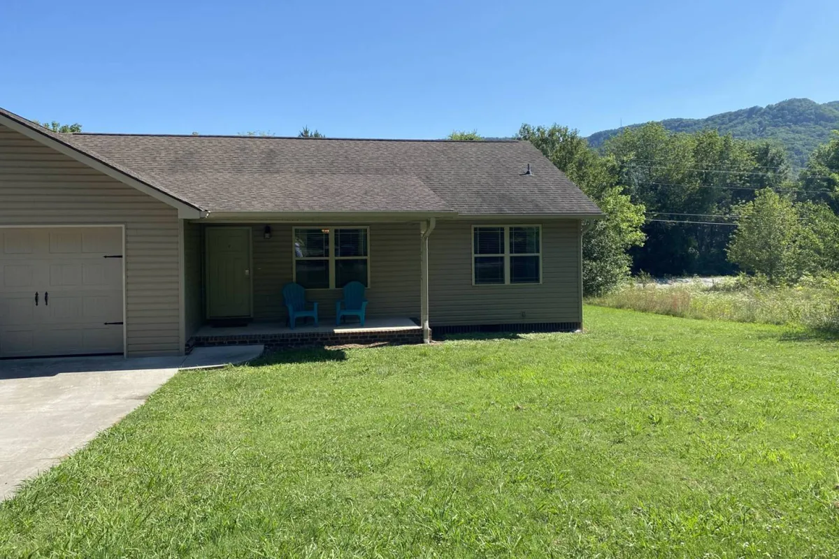 A welcoming exterior view featuring a landscaped lawn, a green front door, and a front porch with blue chairs. The driveway and visible lawn imply available, convenient off-street parking and easy group coordination between the two homes.