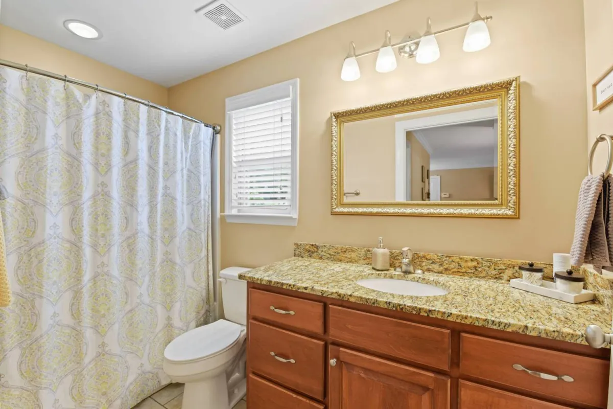 A modern bathroom with a granite countertop, a large framed mirror, and a decorative shower curtain. Sunlight filters in, highlighting plush towels and a serene, well-stocked space designed for comfort and refreshment.