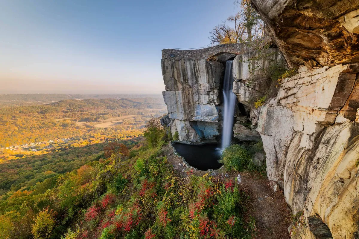 A stunning waterfall cascades over rock formations surrounded by autumn foliage, highlighting the region’s natural beauty. The scene is perfect for hiking, photography, and connecting with nature, alluding to nearby attractions like Lookout Mountain and Raccoon Caverns.