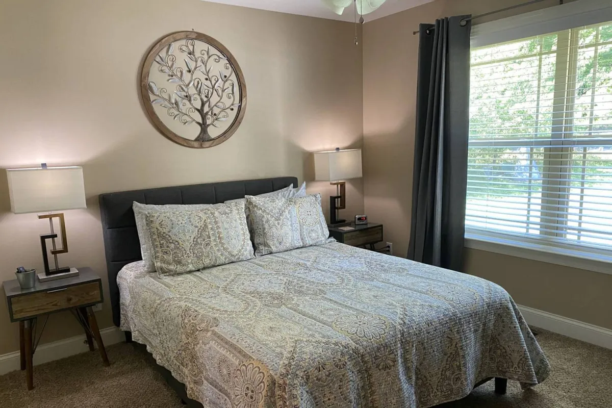 A stylish bedroom featuring an elegantly made bed with decorative wall art, twin nightstands with lamps, and a window allowing soft natural light. The mood conveys comfort and tranquility, perfect for relaxation or unwinding after the day.